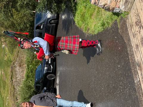       Bagpiper dressed in bright traditional tartan attire playing beside a rural road with parked cars nearby.
  
