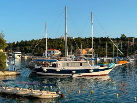      Elegant two-masted yacht moored in picturesque harbor at golden hour with pastel houses and tree-lined hills.
  