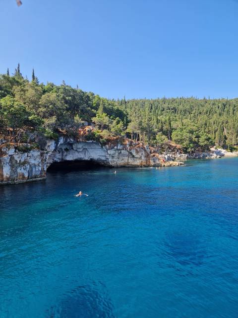       Swimmer enjoying vivid turquoise water near dramatic limestone cave and forested cliffs.
  