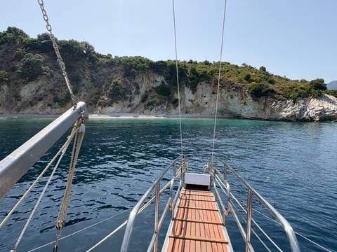       View from sailboat bow toward secluded rocky cove with clear water and green hillside.
  