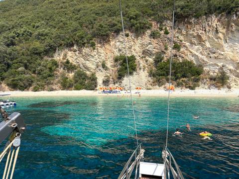       Crystal-clear bay with swimmers near pebble beach, colorful umbrellas and forested cliff backdrop seen from boat rigging.
  