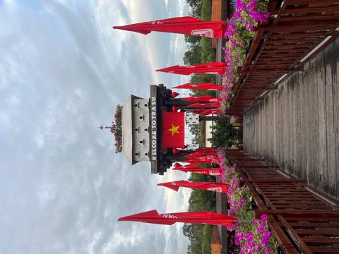       Wooden bridge lined with red flags leads to a white gate tower reading 'Welcome to Tra Su'.
  