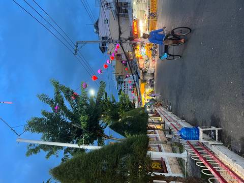       Evening street market decorated with colorful lanterns where locals walk and cycle past food stalls.
  