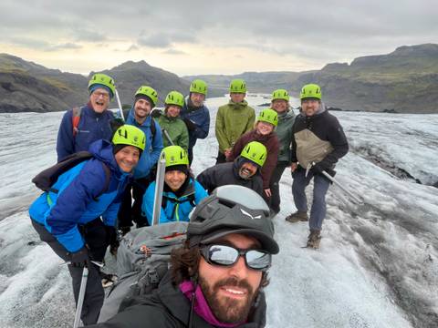       A smiling group of helmeted trekkers poses on a grey glacier with rugged mountains behind them.
  