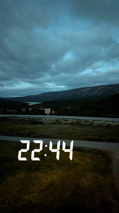       Dim pre-dawn view of a highway and distant lake against dark rolling mountains under a cloudy sky.
  