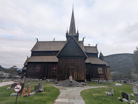       Historic dark-wood stave church stands amid gravestones and mountains while a visitor looks on.
  
