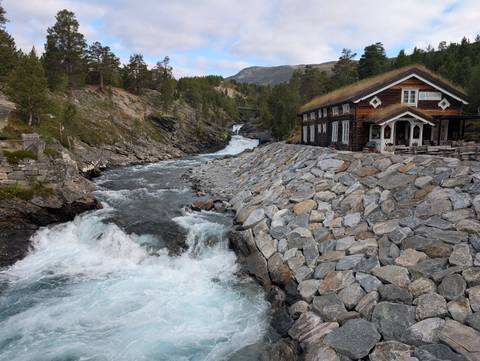       Rushing turquoise river flows past a stone-lined bank and a turf-roof cabin in a mountain valley.
  