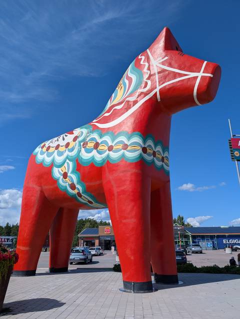       Enormous bright-red painted Dala horse statue stands against a clear blue Scandinavian sky.
  
