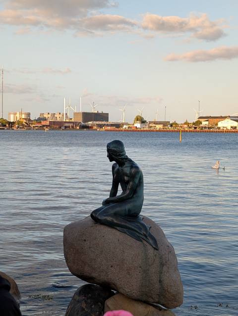       Iconic Little Mermaid statue gazes over the harbor with industrial skyline and wind turbines behind.
  
