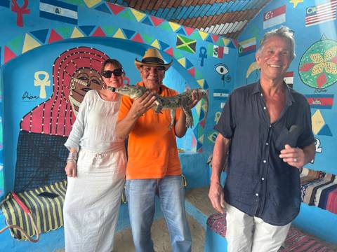       Three travellers indoors hold a small crocodile in a brightly painted Nubian-style room decorated with colourful symbols.
  