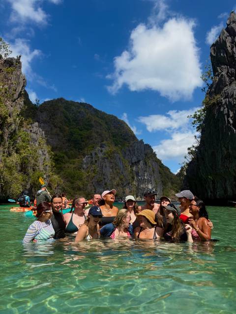       Laughing group of travellers swimming and kayaking in a turquoise lagoon surrounded by towering limestone cliffs in Palawan.
  