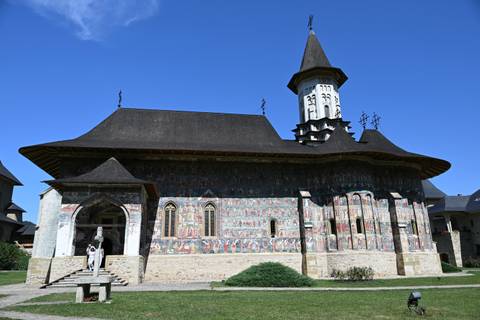       Voroneț Monastery with dark shingled roof and colourful exterior fresco panels under a blue sky.
  