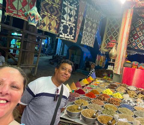       Tourists smile in front of a colorful spice and herb stall at a bustling market.
  