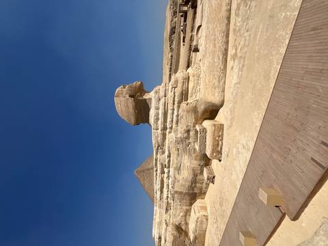       Iconic side profile of the Great Sphinx with the Pyramid of Khafre in the background under a clear sky.
  