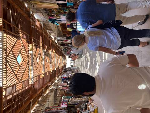       Tourists wander through a sun-patterned covered bazaar lined with textiles and souvenirs.
  