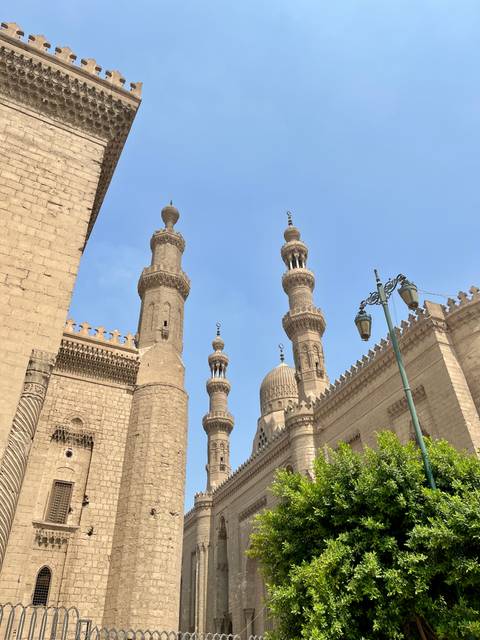       Upward view of ornate stone minarets and domes of a historic Cairo mosque against blue sky.
  