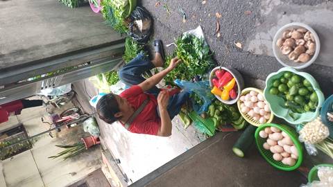       Street vendor sits amid baskets of fresh produce preparing greens in a narrow alley market.
  