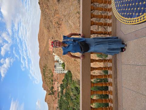       Traveler poses on a balcony overlooking ochre cliffs and a hillside village.
  