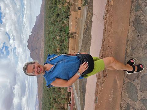       Smiling traveler stands before lush oasis of palm trees and a distant rocky peak.
  