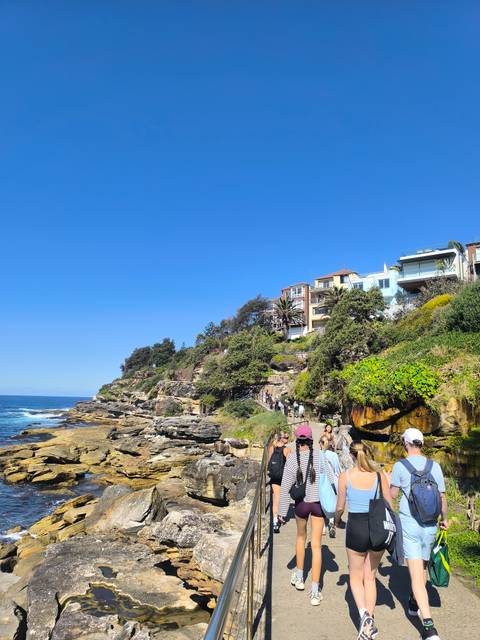       Coastal cliff pathway with walkers and colorful hillside homes overlooking the bright blue sea.
  