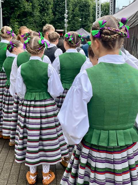       Back view of participants wearing bright green traditional folk vests over striped skirts and white shirts.
  