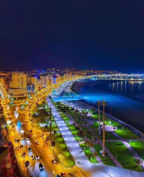      Nighttime panorama of a Moroccan coastal boulevard with bright city lights curving along the shoreline and high-rise buildings overlooking the sea.
  