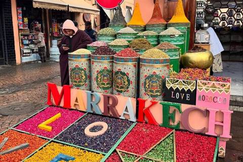       Street stall with piles of colourful spices and a bright block letter sign spelling MARRAKECH while a hooded woman checks her phone.
  
