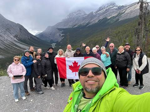       Large tour group poses with Canadian flag for a selfie in front of dramatic Rocky Mountain valley.
  