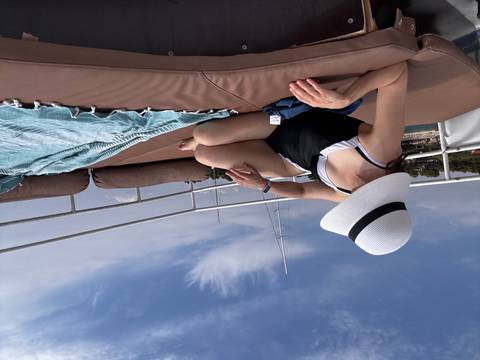       Reclining woman in swimwear relaxes on a cushioned deck of a yacht under a bright sky.
  