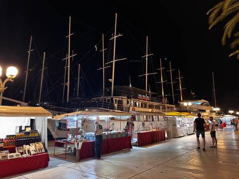       Nighttime market stalls line a marina filled with tall-masted sailing yachts under lamplight.
  