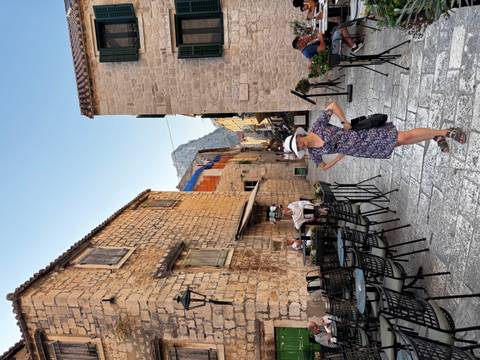       Cobbled alley of a stone coastal town with café tables; woman in floral dress stands posing.
  