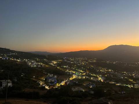      Twilight panorama of a hilly Moroccan town with lights twinkling beneath an orange sky.
  
