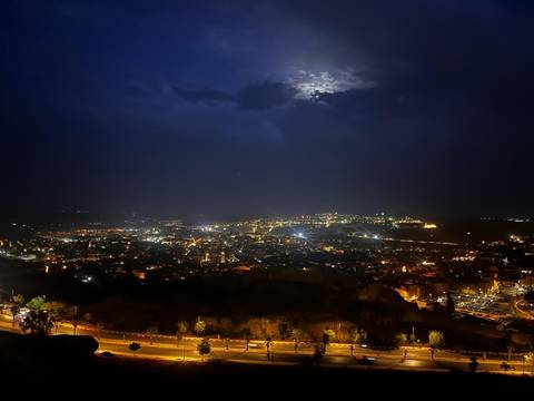       Night panorama of a sprawling city lit by streetlights under a cloudy moonlit sky.
  