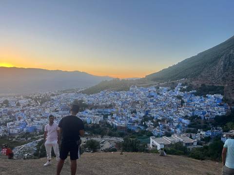       Overlook of Chefchaouen’s blue houses at sunset with visitors enjoying the view in the foreground.
  