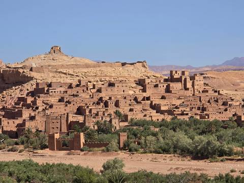       The earthen village of Ait Benhaddou sits atop a desert hill surrounded by sparse greenery beneath a clear sky.
  