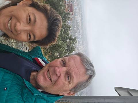       Close selfie of two travelers on a misty hilltop overlooking a faint city panorama.
  