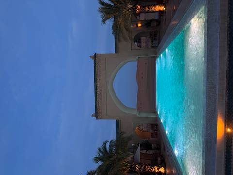       An illuminated infinity pool under an arched gateway looks onto silent night-time desert dunes.
  