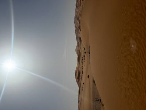       Midday sun streams over sweeping Sahara dunes, a bright flare cutting the clear sky.
  