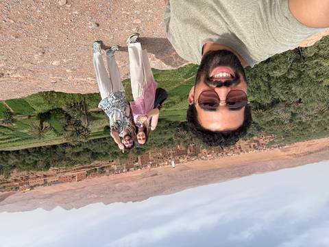       Three smiling travelers capture a selfie overlooking a fertile oasis and adobe village backed by barren mountains.
  