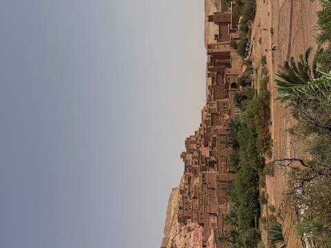       The earth-toned ramparts of Ait Benhaddou stand against a pale sky above sparse desert brush.
  
