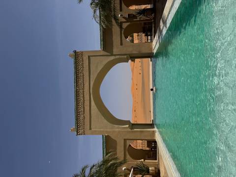       A person floats peacefully in a turquoise pool framed by Moorish arches with open desert beyond.
  