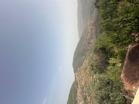       Expansive mountain vista with green valleys and reddish cliffs under a hazy blue sky
  