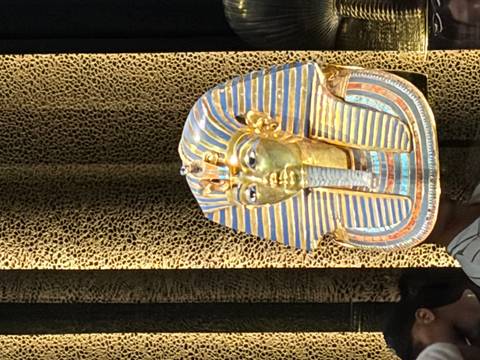       Golden funerary mask of Tutankhamun displayed in a museum case with patterned backdrop.
  