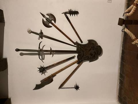       Indoor museum display of medieval weapons arranged around a decorative shield on a white plaster wall.
  