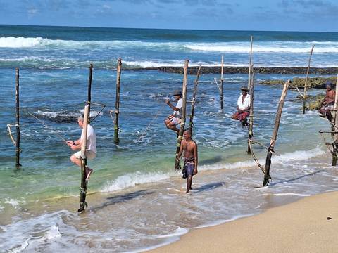      Traditional stilt fishermen balance on poles above turquoise surf along a tropical shoreline.
  
