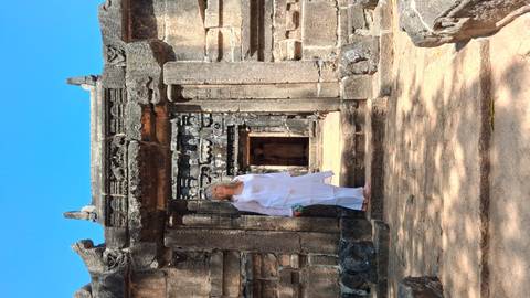       Woman in white attire stands at the stone entrance of an ancient ruin complex under bright blue sky.
  