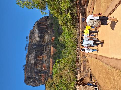       Line of tourists walking along a sandy path toward the massive vertical face of Sigiriya Rock.
  