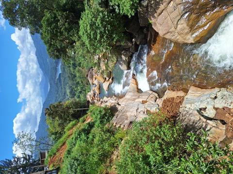       Mountain stream cascading over smooth rocks through lush green valley with distant lakes and peaks under blue sky.
  