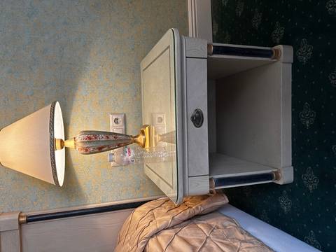       Bedside table with classic lamp, bottled water and empty shelf below in a dated hotel room.
  