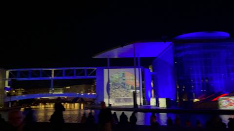       Nighttime water projection show illuminating a modern glass building in blue beside the Spree River.
  